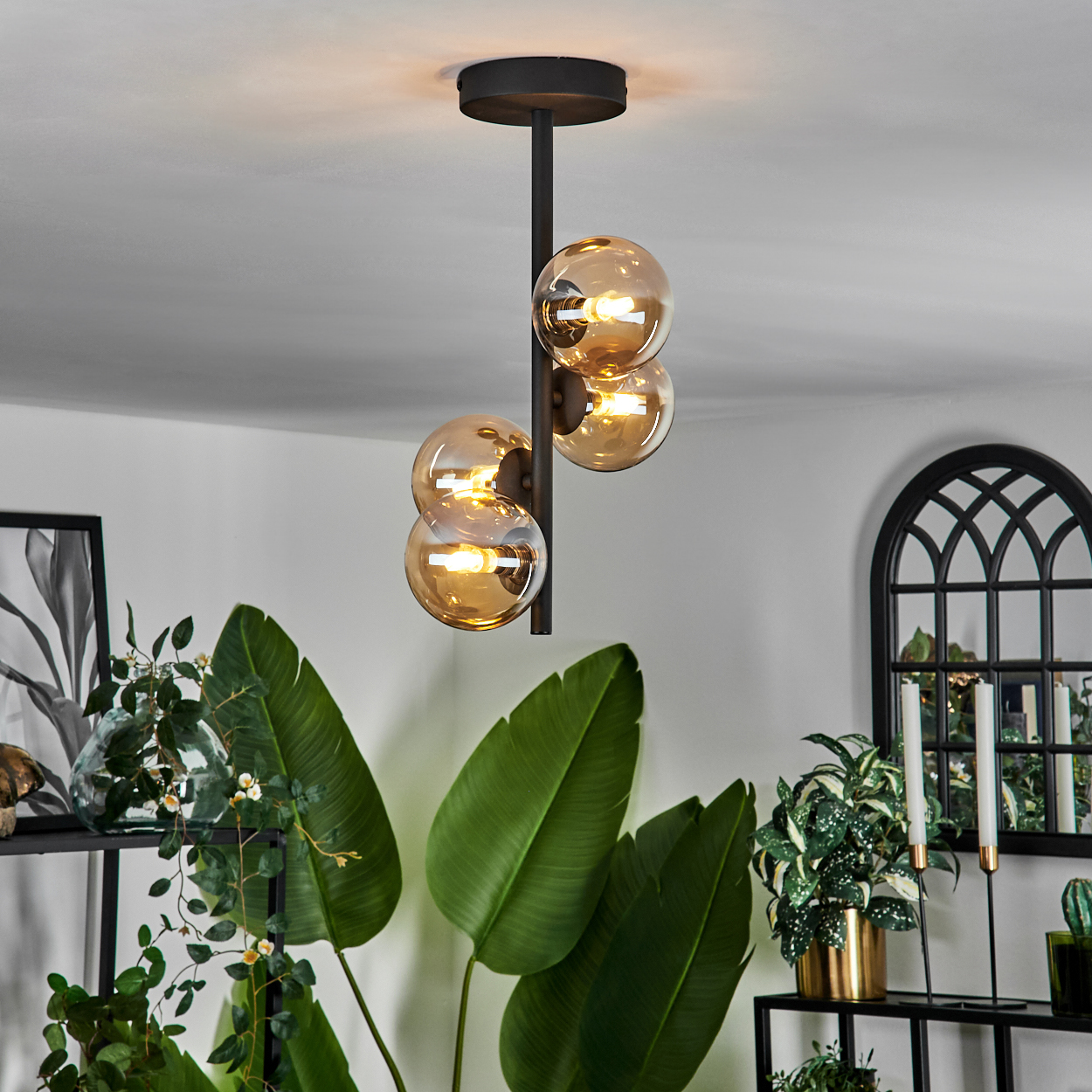 Eine Deckenlampe mit drei orangefarbenen Kugeln. Der Hintergrund umfasst Pflanzen, ein Fenster und einen schwarzen Bogen.