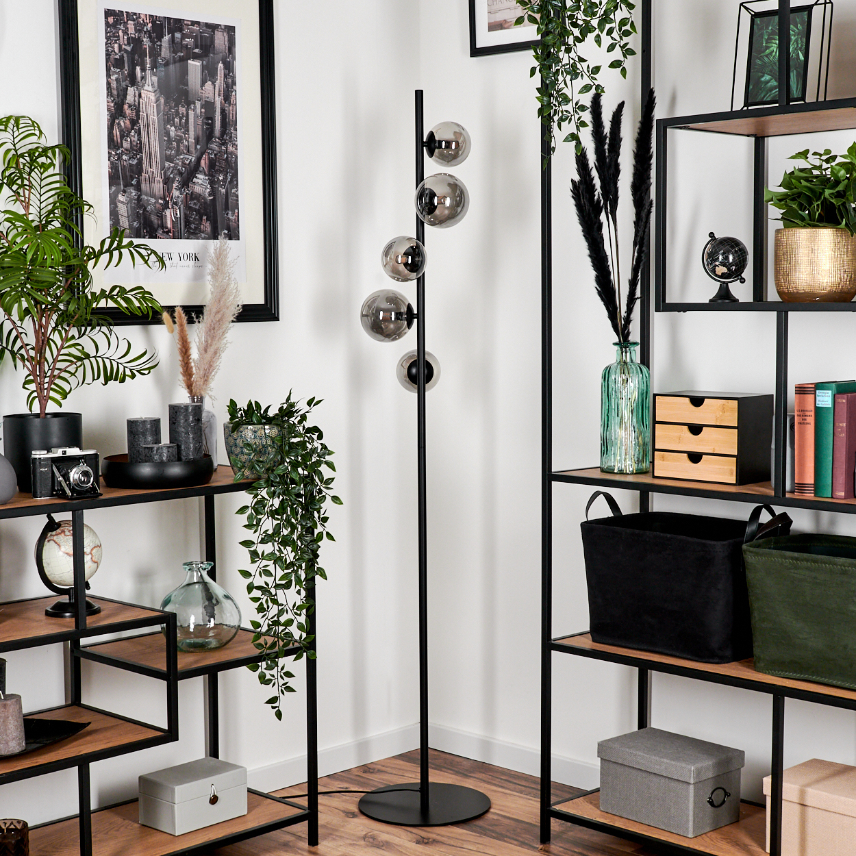 Ein Raum mit Regalen, Pflanzen und einer schwarzen Stehlampe. Die Lampe hat fünf graue Glaskugeln, mit New York im Hintergrund.