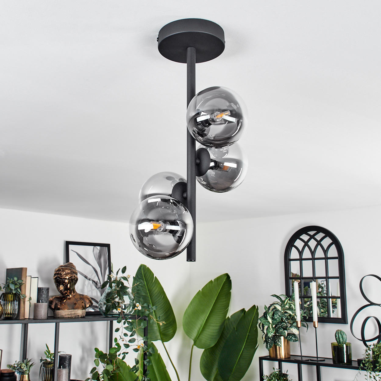 Eine Deckenlampe mit vier Glaskugeln. Die Glühbirnen sind an. Ein Raum mit Pflanzen, Kunst und Möbeln ist im Hintergrund.