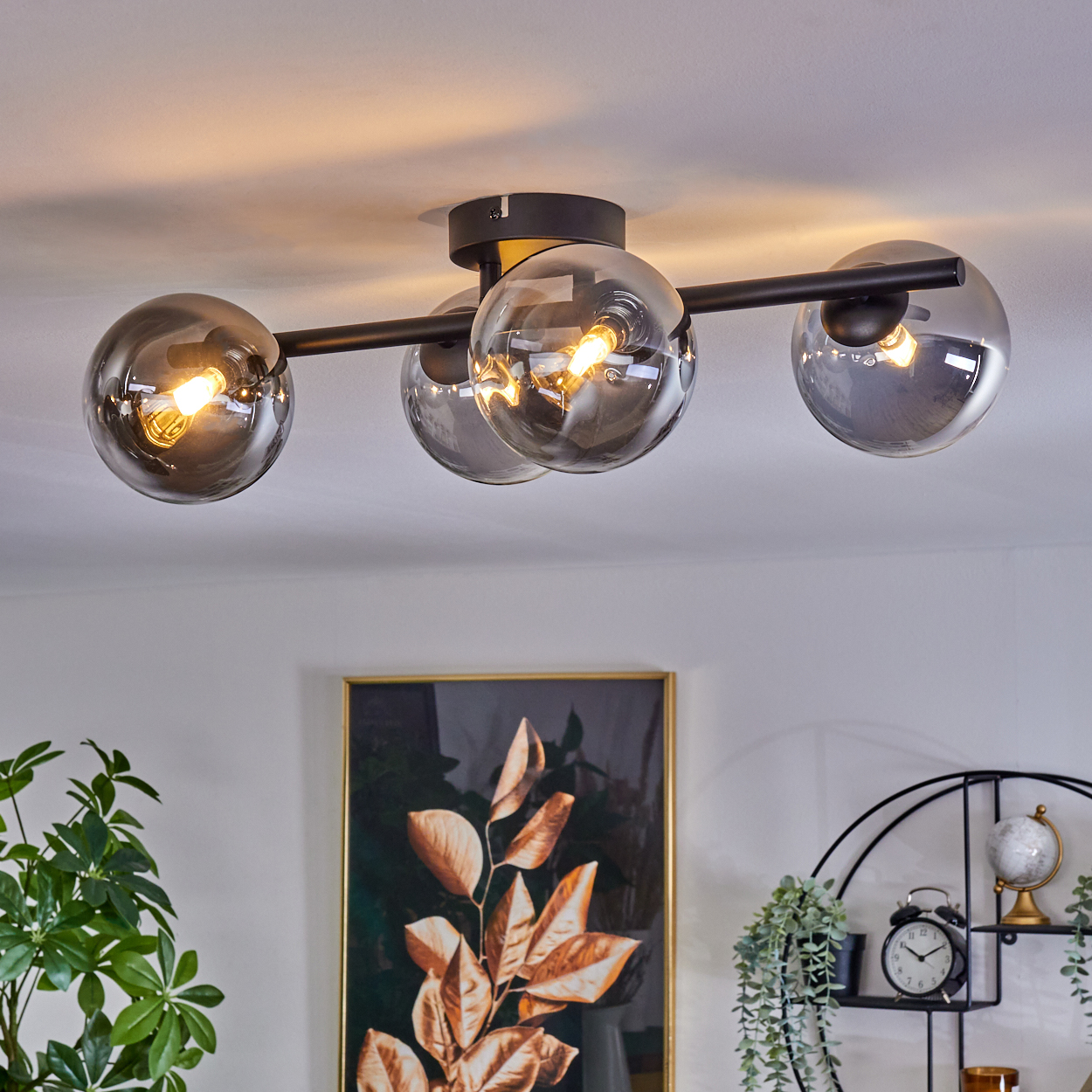 Deckenlampe mit vier Glaskugeln. Schwarzer Rahmen. Grüne Pflanzen und ein Gemälde im Hintergrund.