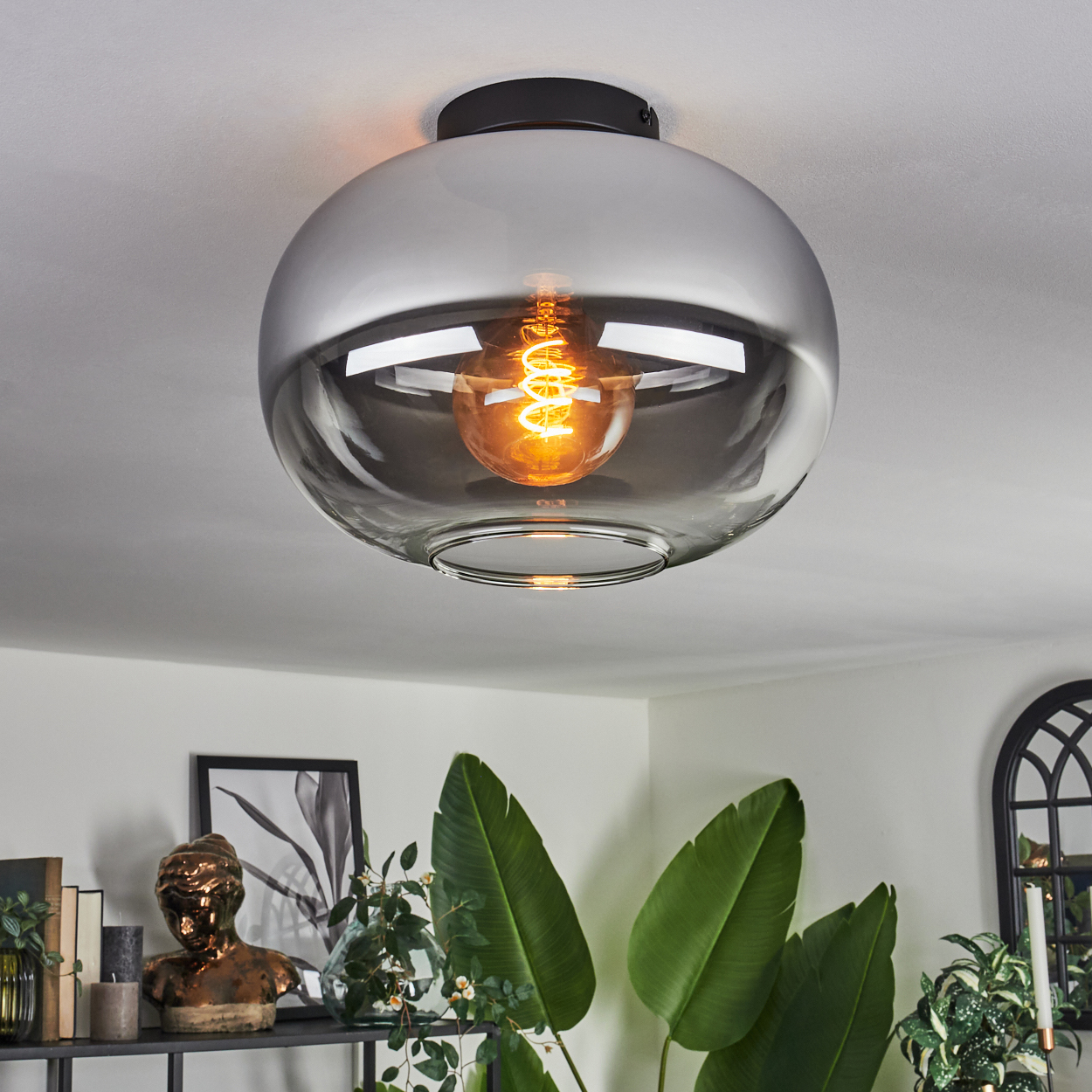 Eine runde, graue Glas-Deckenlampe mit sichtbarer Glühbirne hängt von einer weißen Decke ab, Blätter unten.