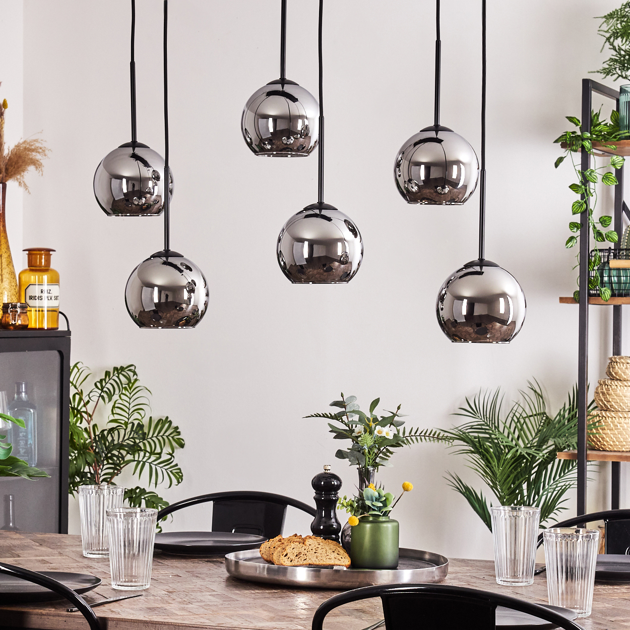 Sechs silberne Kugelhängeleuchten über einem Holztisch mit Tellern, Gläsern und Mittelstücken. Pflanzen sind sichtbar.