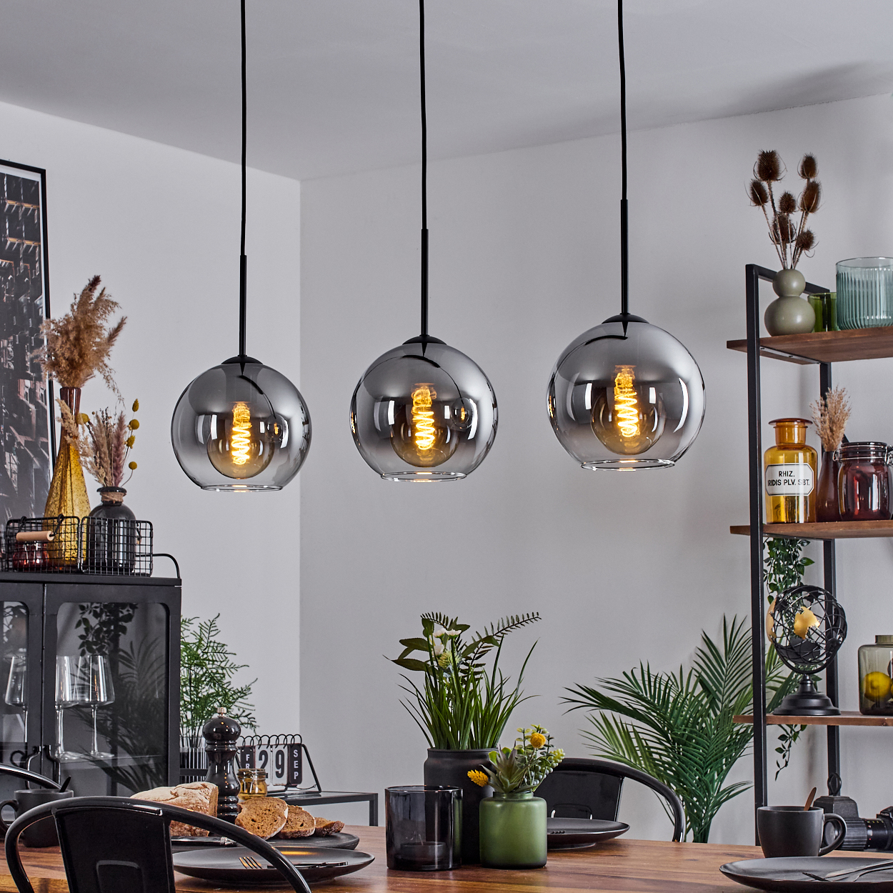 Drei Pendelleuchten mit kugelförmigen Glasschirmen, jede mit einer sichtbaren Glühbirne, hängen über einem Holztisch.