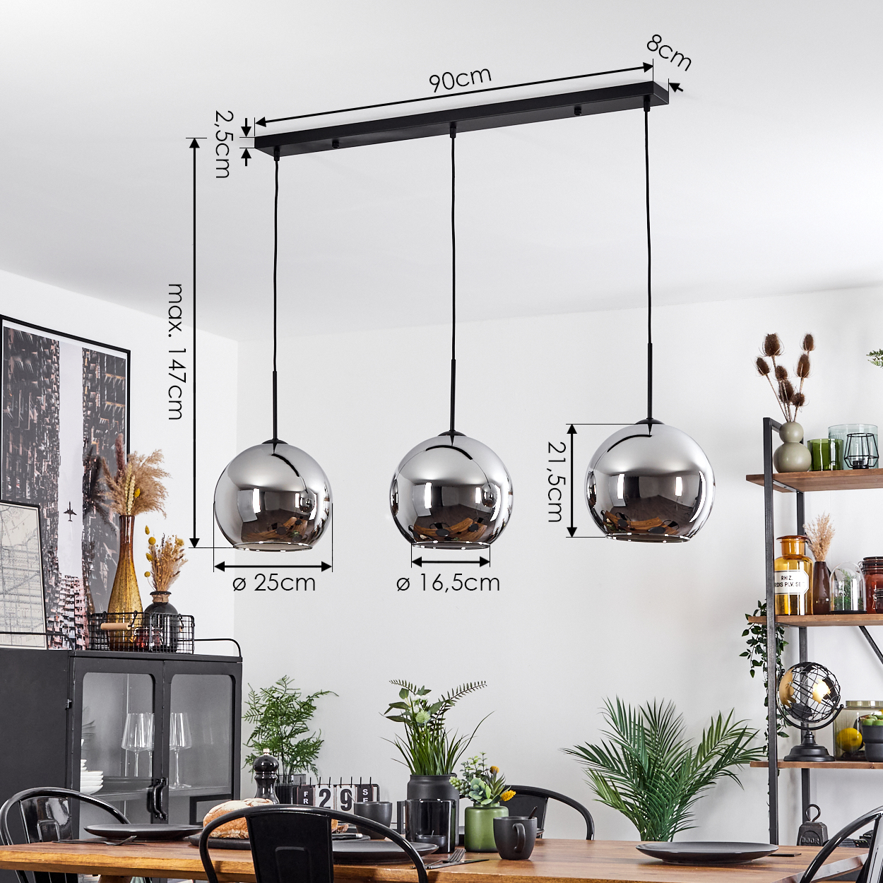 Hängelampe mit 3 metallischen Kugeln. Die Kugeln hängen über einem Holztisch. Ein Regal und ein Schrank sind im Hintergrund.