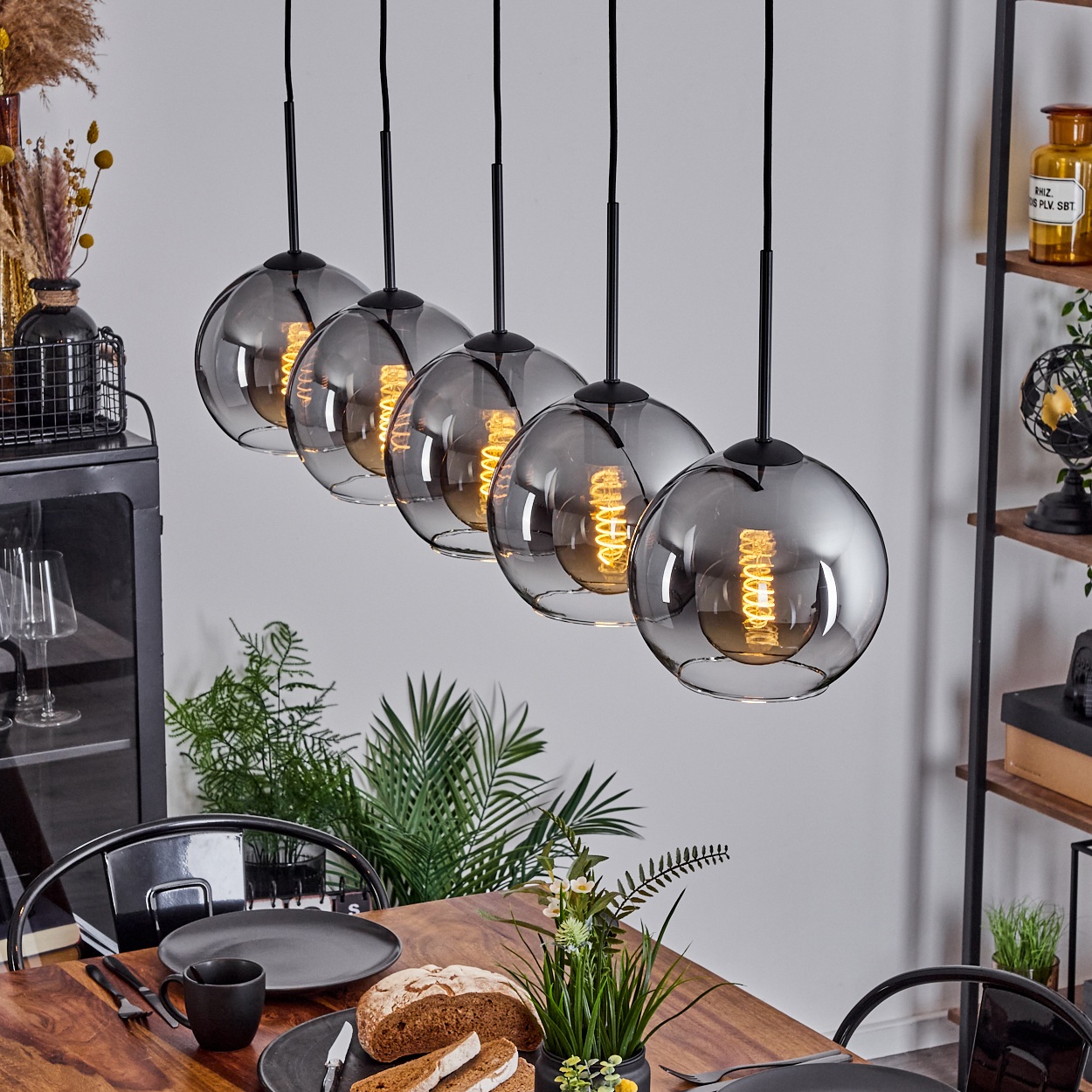 Sechs Glas-Pendelleuchten mit leuchtenden Glühbirnen hängen über einem Holztisch mit Essen. Andere Dekoration ist sichtbar.
