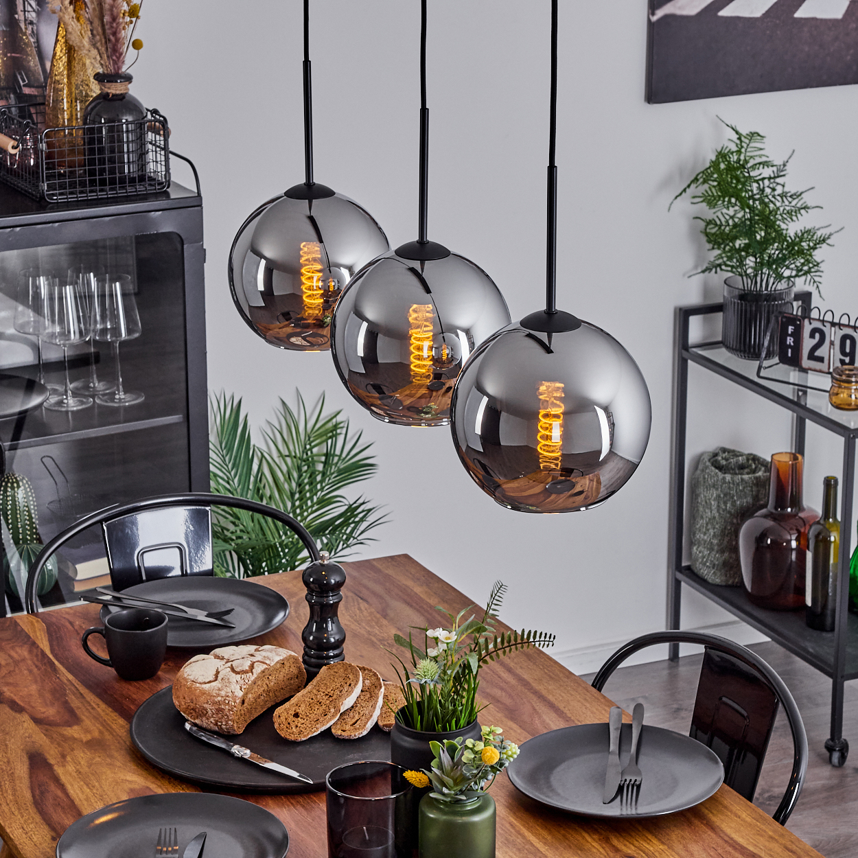 Esszimmer mit drei kugelförmigen Pendelleuchten. Holztisch mit Essen, Pflanzen und Dekorationen. Dunkle Wände.