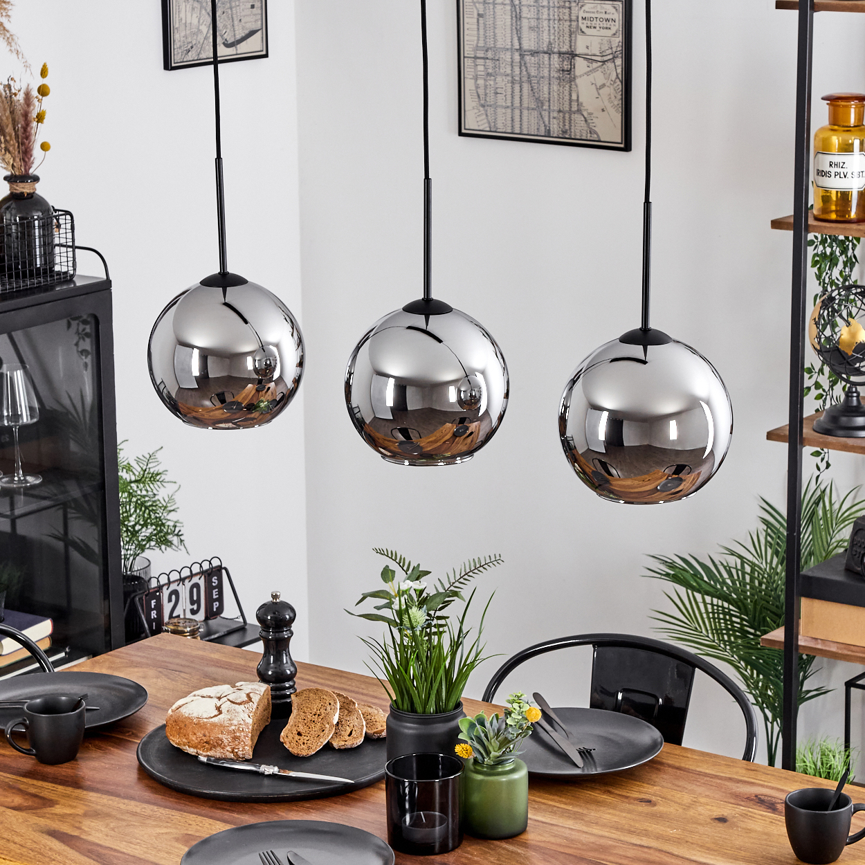 Esszimmer mit drei silbernen Pendelleuchten. Holztisch, schwarze Teller und Dekorationen. Der Raum ist gut beleuchtet.
