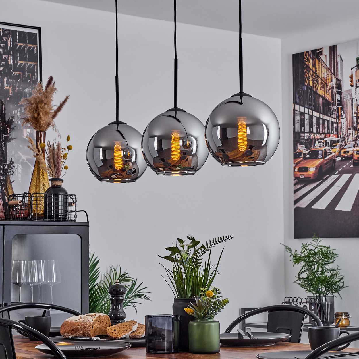 Esszimmer mit drei Pendelleuchten. Holztisch mit Essen und Dekorationen. Ein Stadtbildgemälde an der Wand.