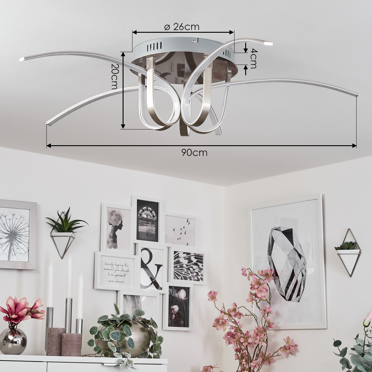 Eine Deckenlampe mit geschwungenen Armen, Maße werden angezeigt. Der Hintergrund ist eine weiße Wand mit Kunst und Deko.