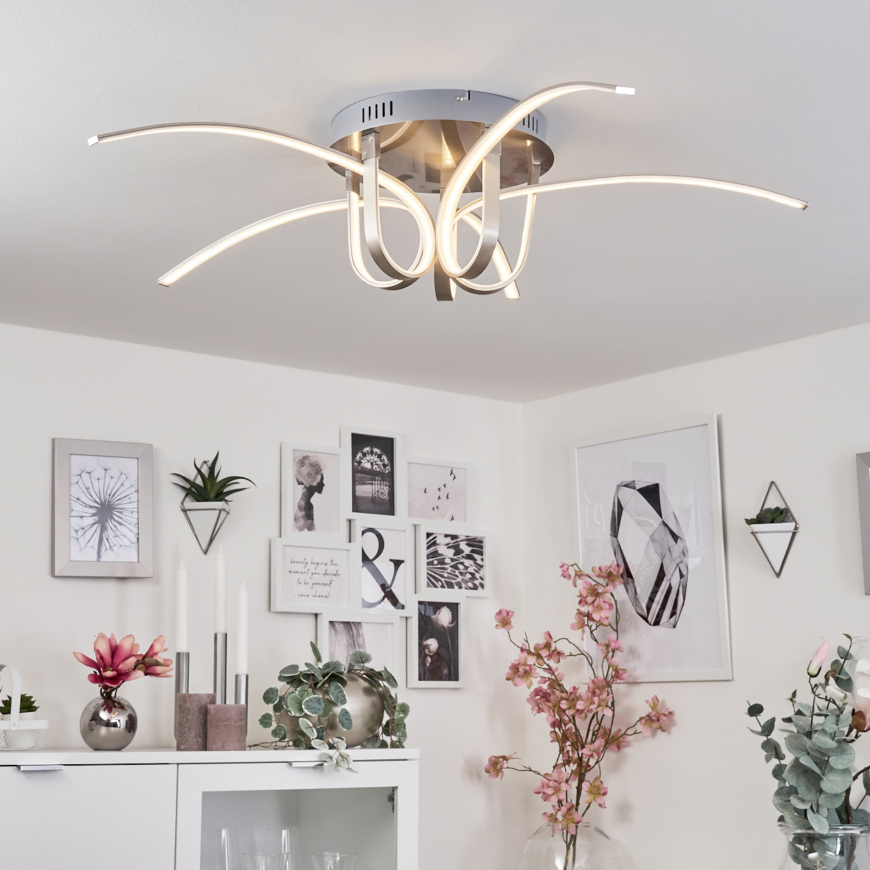 Eine beleuchtete Deckenlampe mit gebogenen silbernen Armen. Hintergrund: weiße Wand mit dekorativen Bildern und Blumenvase.