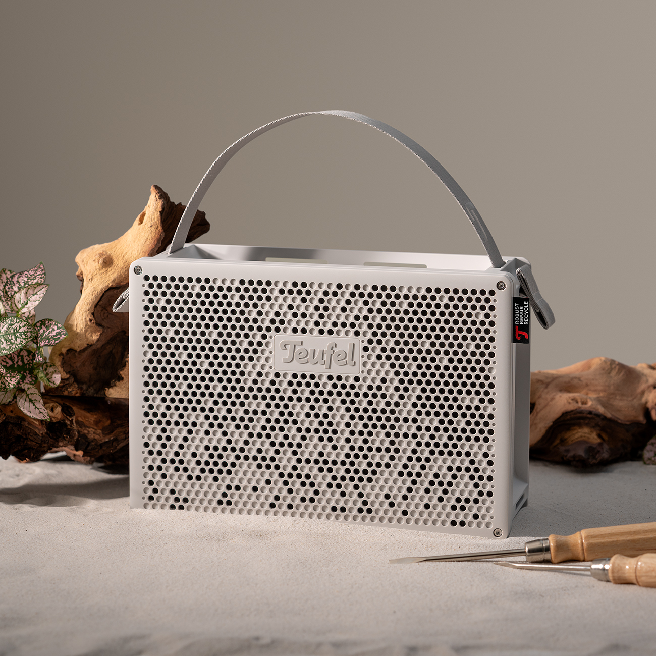 Haut-parleur blanc avec poignée, logo Teufel, sur sable. Bois et outils en arrière-plan. Étiquette rouge.