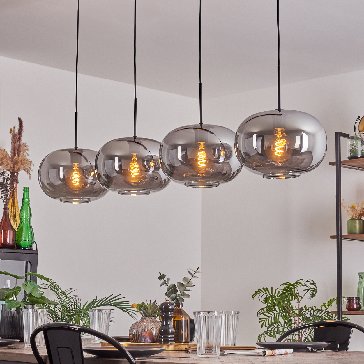 Vier Pendelleuchten mit silbernen Glaskugelschirmen, beleuchtet, hängen über einem Tisch. Hintergrund umfasst Deko und Tisch.