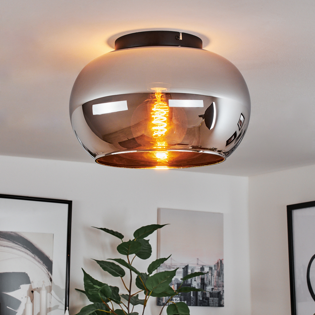 Eine verchromte Deckenlampe mit einer leuchtenden Glühbirne. Vor einer weißen Decke, mit Pflanze.