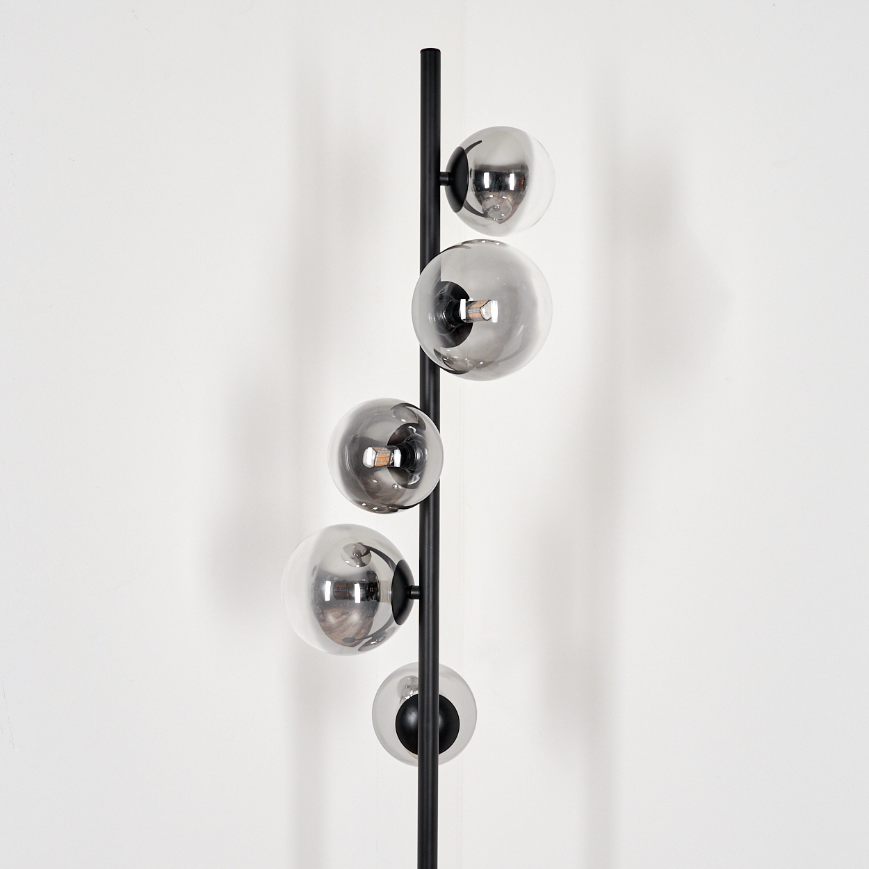 Eine Stehlampe mit 5 kugelförmigen Glasschirmen. Die Lampe steht vor einer weißen Wand. Glühbirnen leuchten.