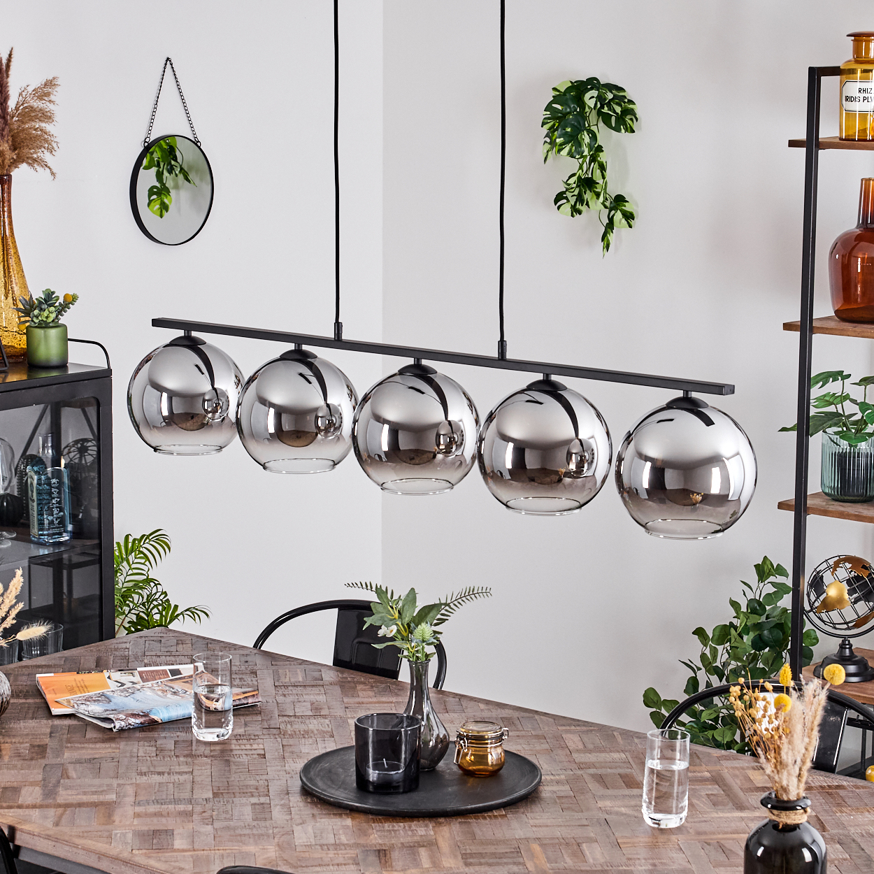 Ein Kronleuchter mit fünf kugelförmigen Glasschirmen über einem Holztisch. Der Raum hat Pflanzen und einen Spiegel an der Wand.