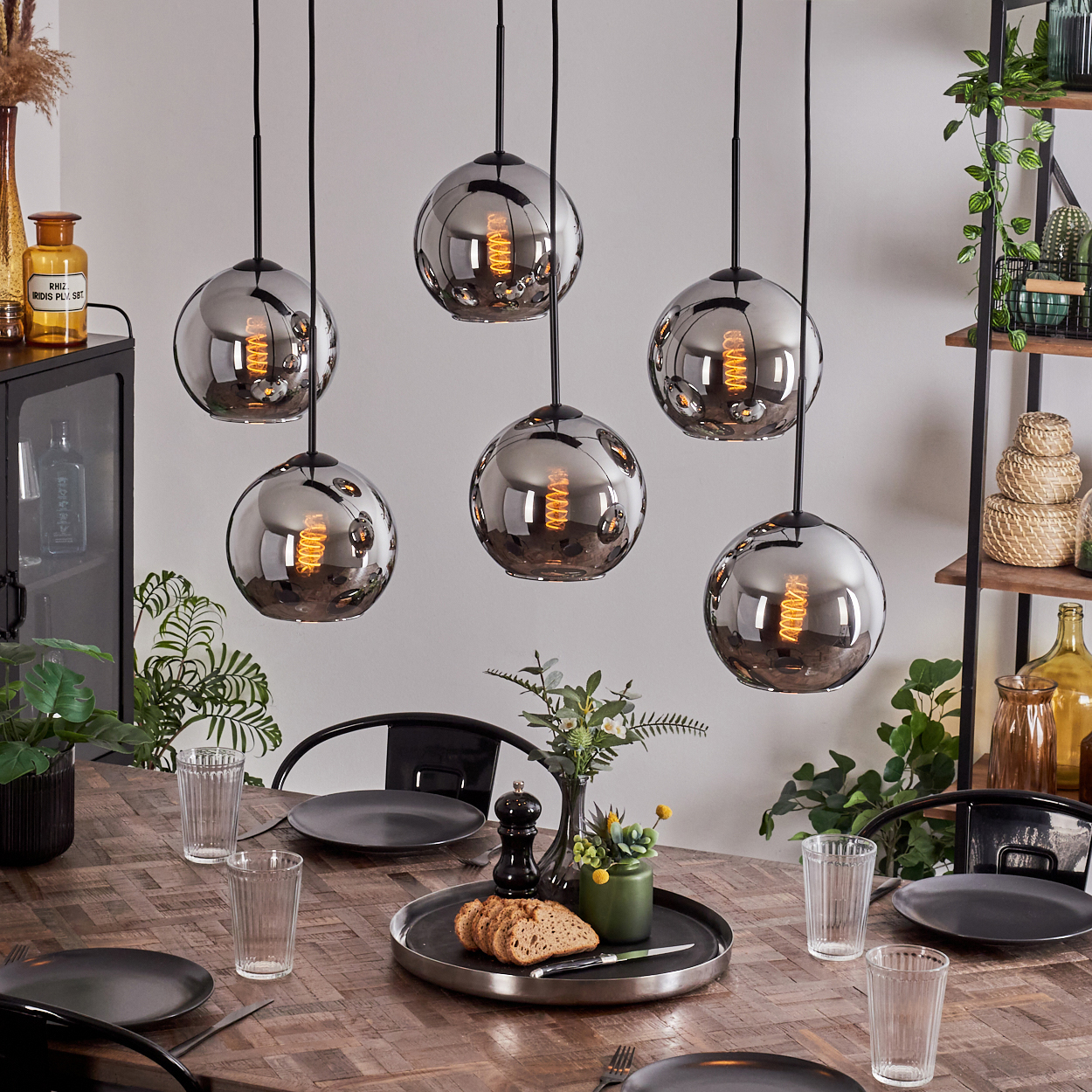 Esszimmer mit Holztisch, sechs hängenden Kugelleuchten und Pflanzen. Glaswaren und ein Tablett mit Brot sind auf dem Tisch.