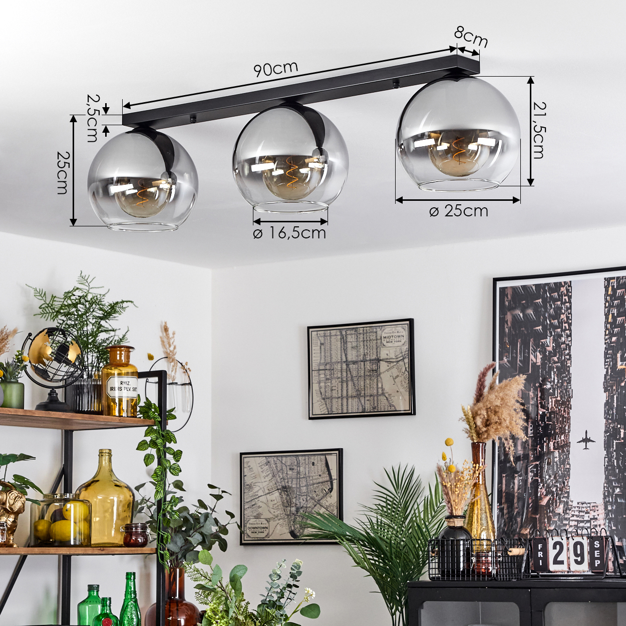 Drei silberne Pendelleuchten hängen von der Decke. Abmessungen sind angegeben. Der Hintergrund umfasst einen Tisch und Kunstwerke.