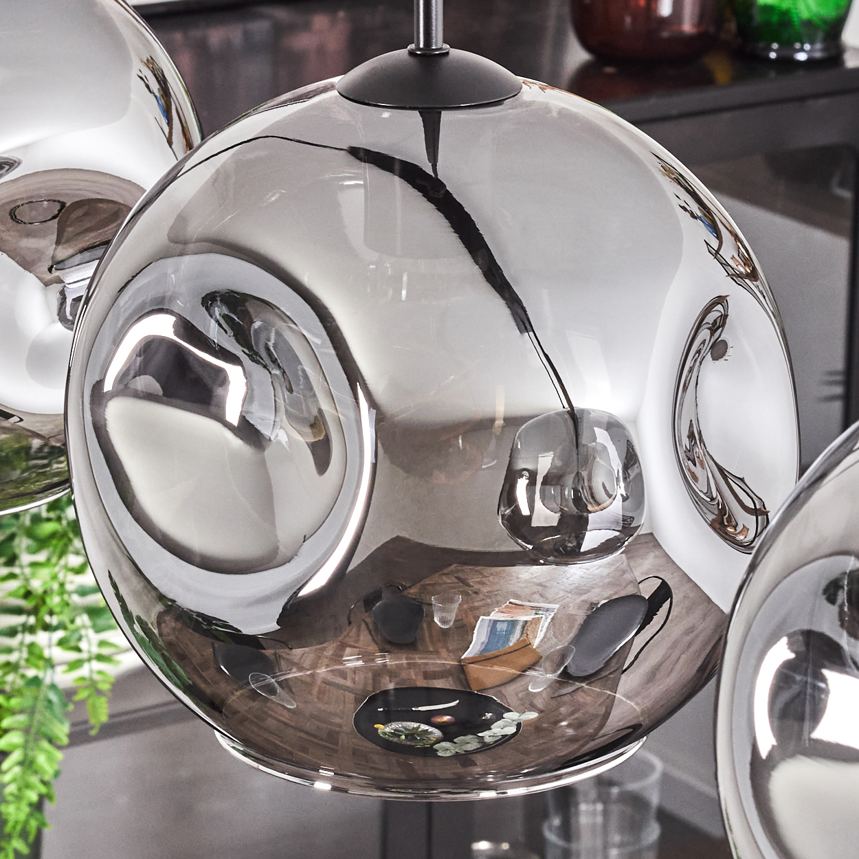 Nahaufnahme einer Pendelleuchte mit grauem Glasschirm. Das Glas reflektiert die Umgebung. Ein Farn ist im Hintergrund.