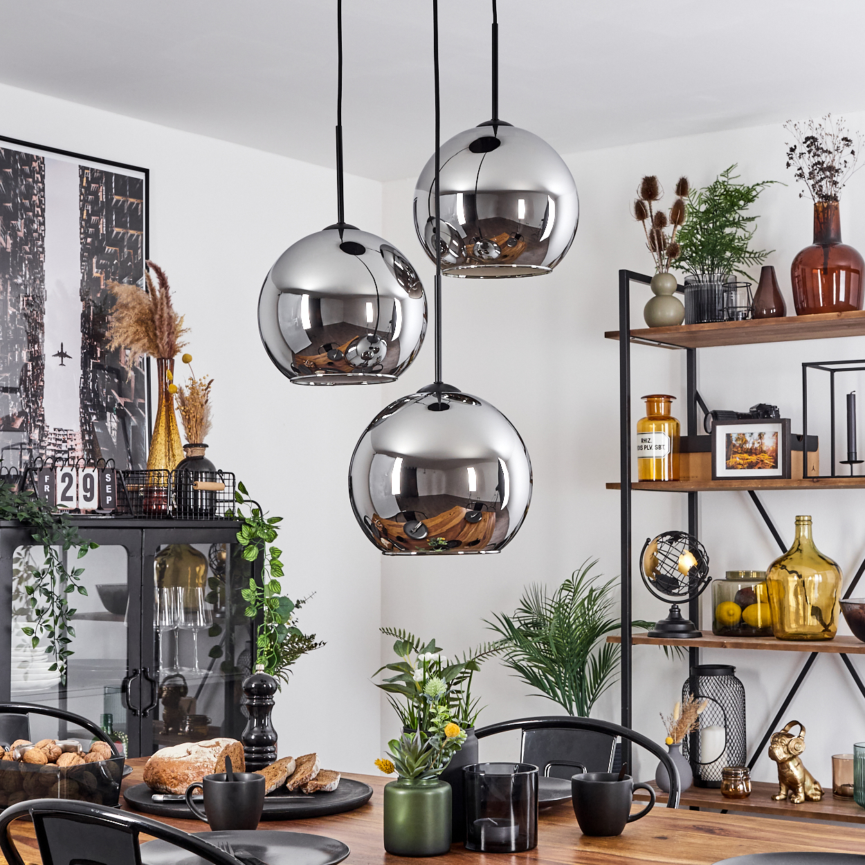 Drei Pendelleuchten hängen über einem Esstisch. Der Raum hat eine moderne Esszimmer-Einrichtung mit Dekorationen und Pflanzen.