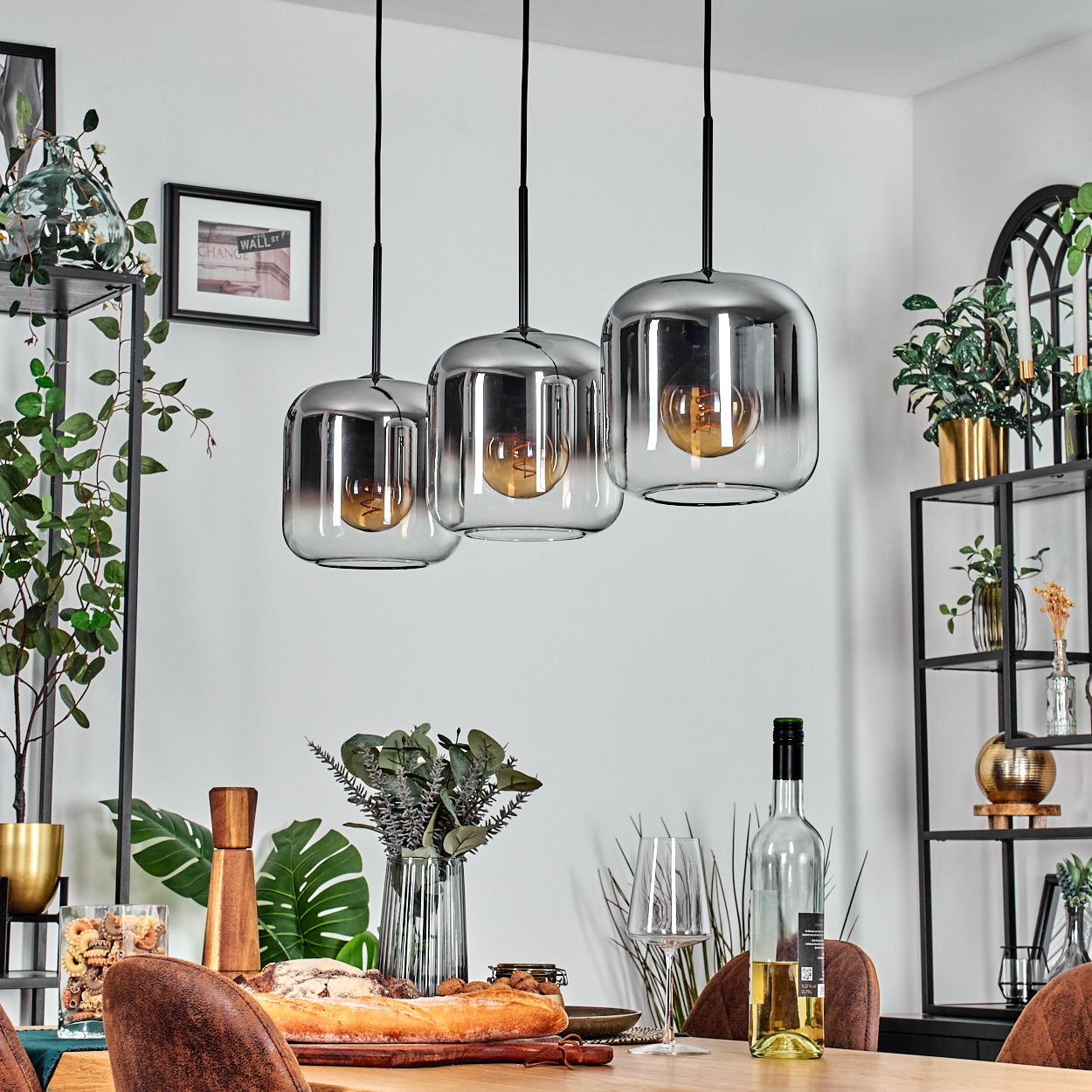 Ein Esszimmer mit Holztisch, drei Pendelleuchten und Zierpflanzen. Eine Weinflasche und ein Glas sind sichtbar.