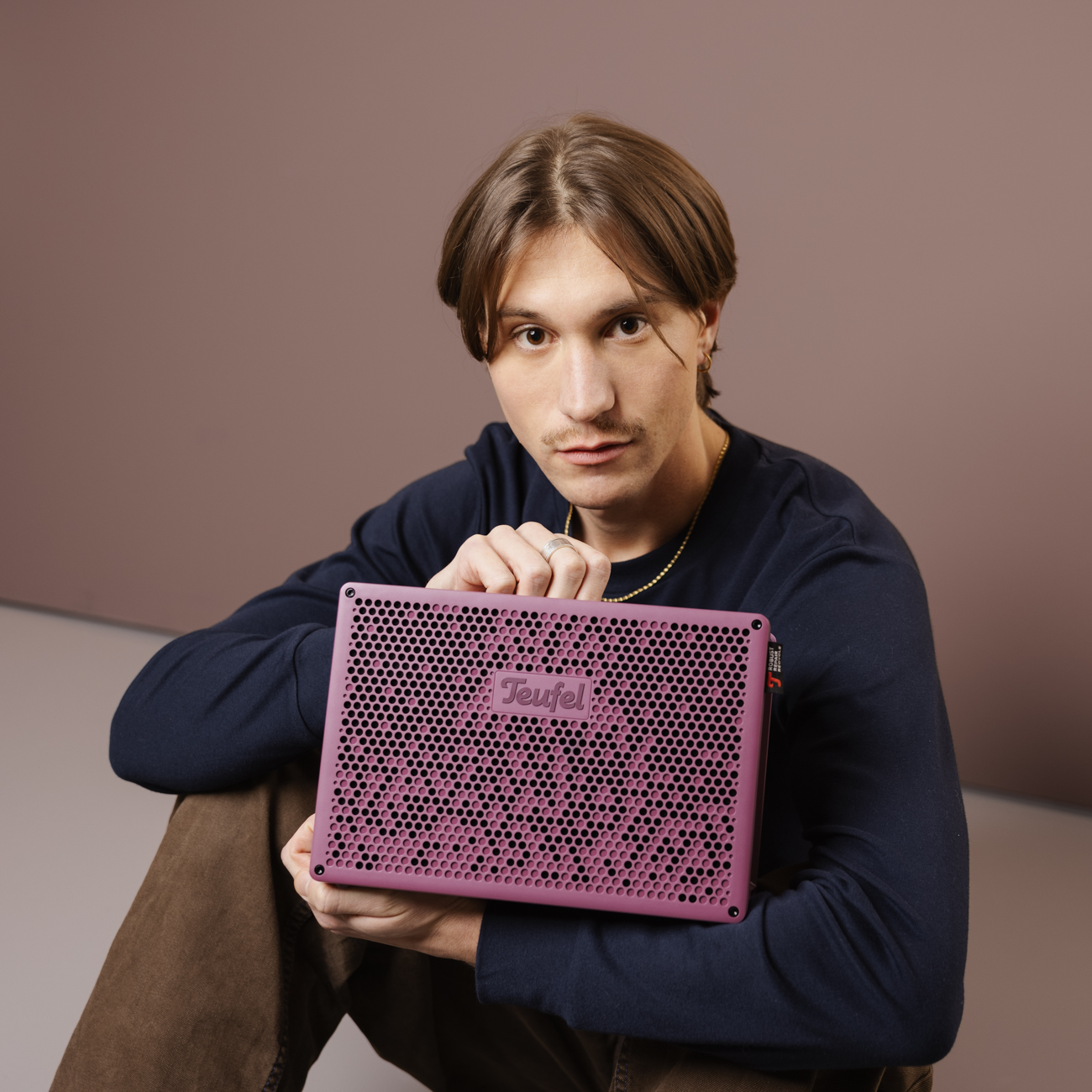 Un homme tient un haut-parleur violet avec un logo. Il est dans une pièce avec un fond neutre.