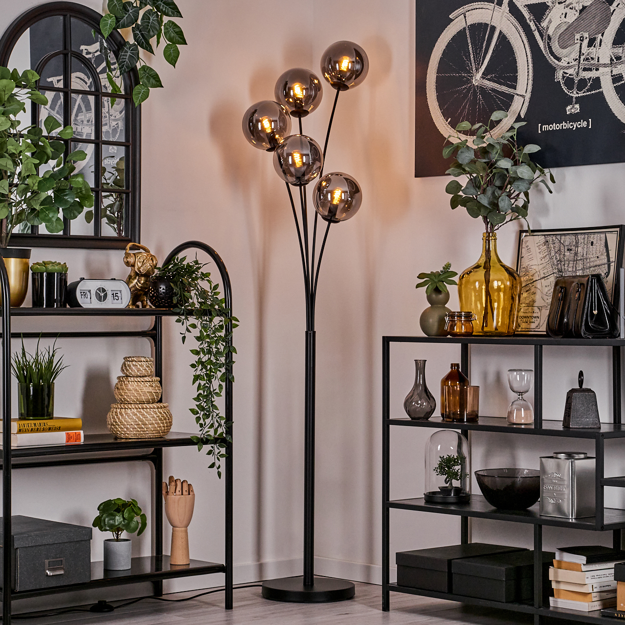 Eine Stehlampe mit fünf kugelförmigen Lampenschirmen, in einem Raum mit Regalen, Kunstwerken und Dekoration. Die Lampe ist an.