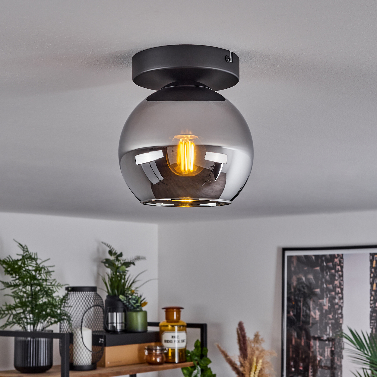 Eine Deckenlampe mit schwarzem Sockel und einer Kugelleuchte. Die Glühbirne ist sichtbar, Raum im Hintergrund.