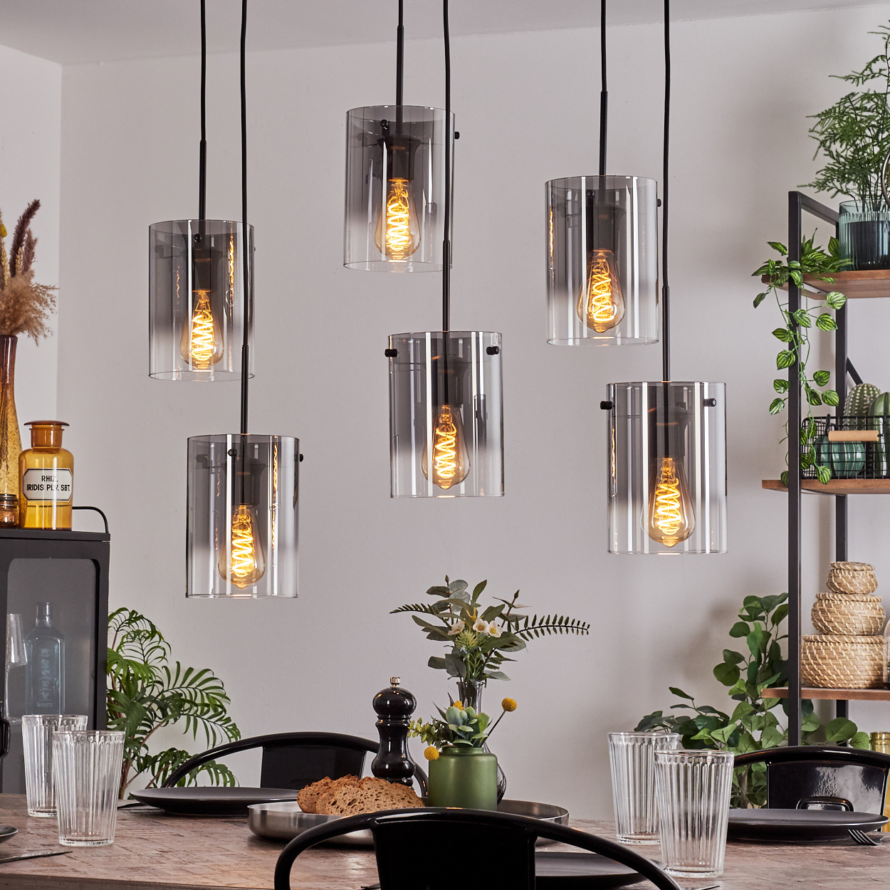 Sechs Hängelampen mit Glaszylinderschirmen und Glühbirnen. Die Lampen befinden sich über einem Holztisch.