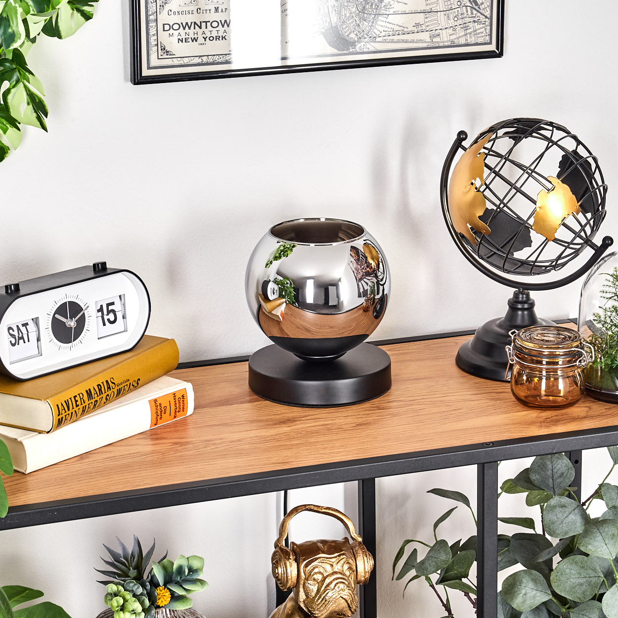 Eine Lampe mit einer verchromten Kugel auf einem schwarzen Sockel, auf einem Regal mit Büchern, einer Uhr und einem Globus.