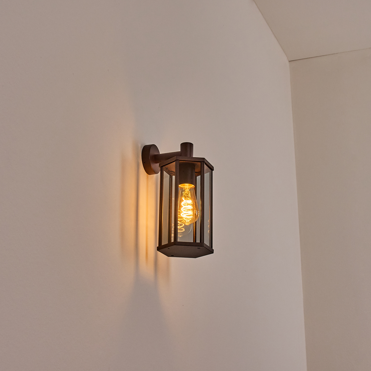 Wandleuchte mit rechteckigem Glasschirm und Wendelglühbirne. Sie ist an einer neutralen Wand befestigt.