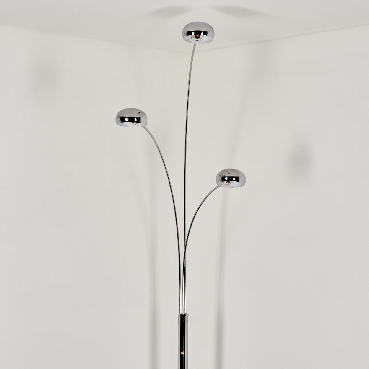 Eine verchromte Stehlampe mit drei kugelförmigen Glühbirnen, in einer Ecke vor einer weißen Wand platziert.