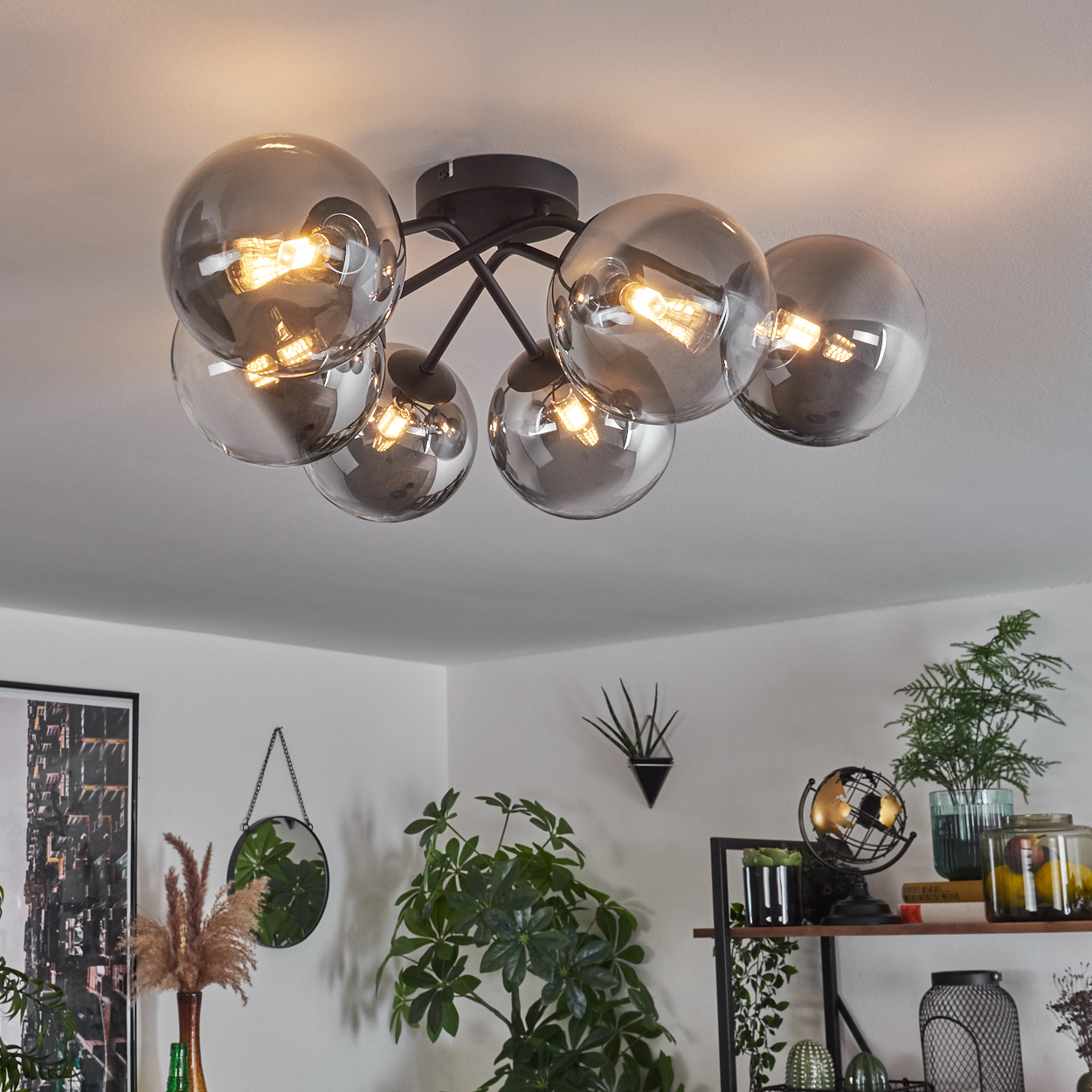 Eine Deckenlampe mit sechs Lichtern und Glühbirnen. Der Raum hat Pflanzen und Regale mit Dekorationen.