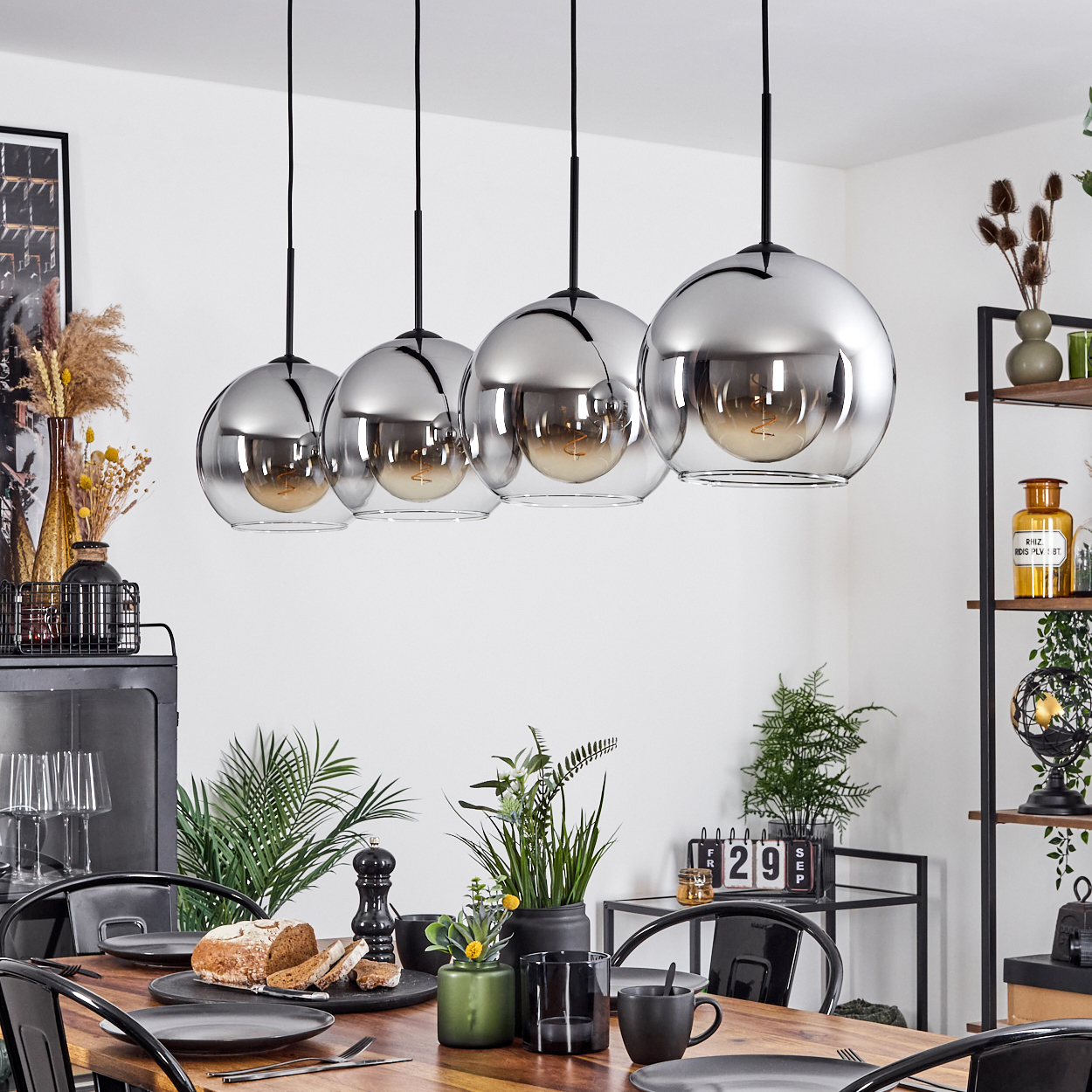 Ein Esszimmer mit Tisch, vier Pendelleuchten und Dekoration wie Pflanzen und einem schwarzen Schrank.