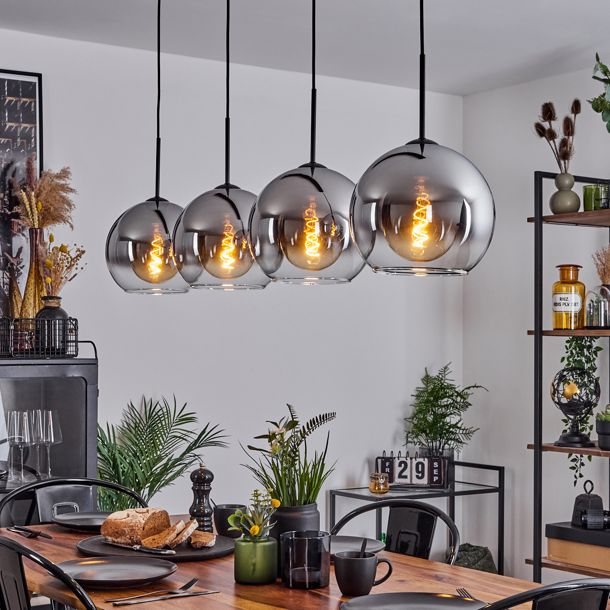 Ein Esszimmer mit Tisch, vier Pendelleuchten und Dekoration wie Pflanzen und einem schwarzen Schrank.