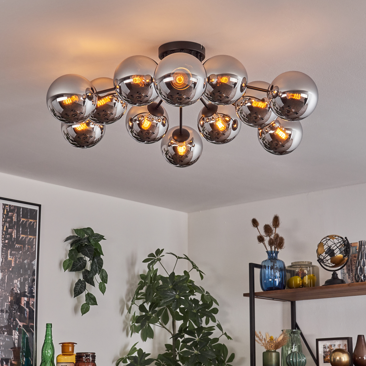 Eine Deckenlampe mit mehreren Chromkugeln und warmen Lichtern. Der Hintergrund zeigt einen Raum mit Pflanzen.