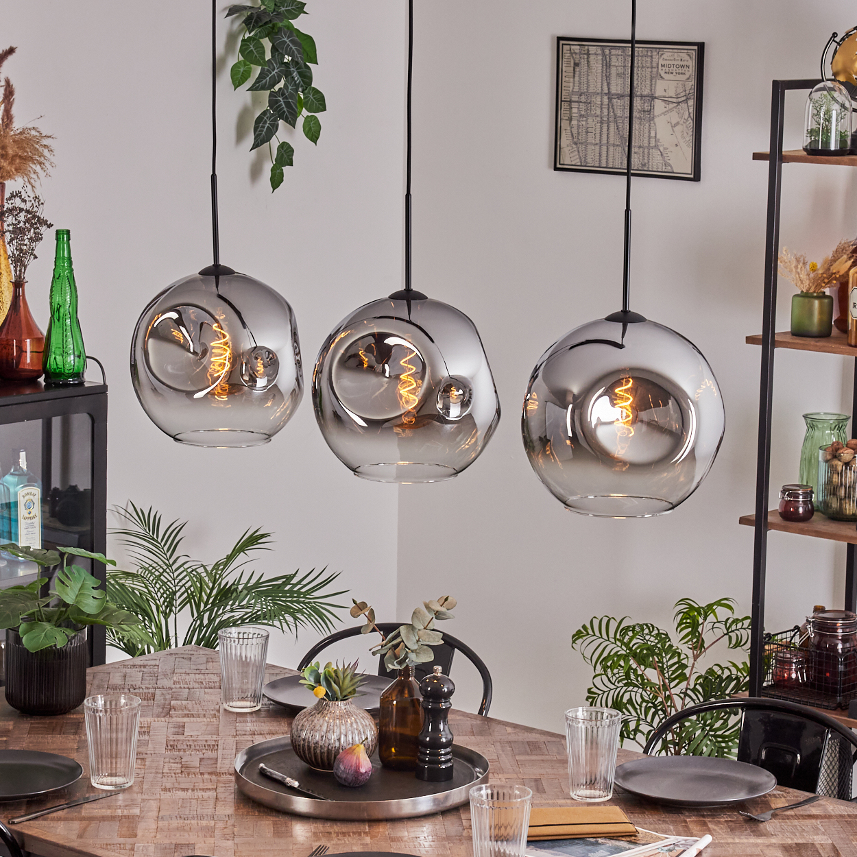Drei graue Glas-Pendelleuchten hängen über einem Holztisch, umgeben von Pflanzen und Dekoration. Jede hat eine sichtbare Glühbirne.