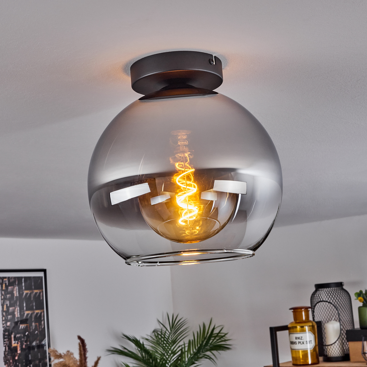 Eine Glaskugel-Deckenleuchte mit dunklem Metallsockel. Die Glühbirne beleuchtet die Kugel, vor einer weißen Decke.