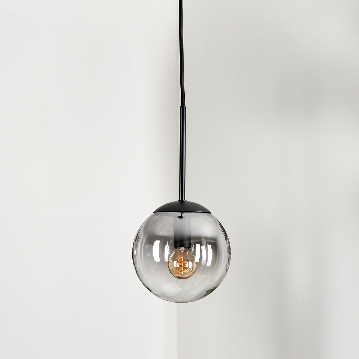 Eine Pendelleuchte mit Glaskugel und sichtbarer Glühbirne hängt von der Decke. Der Hintergrund ist eine weiße Wand.