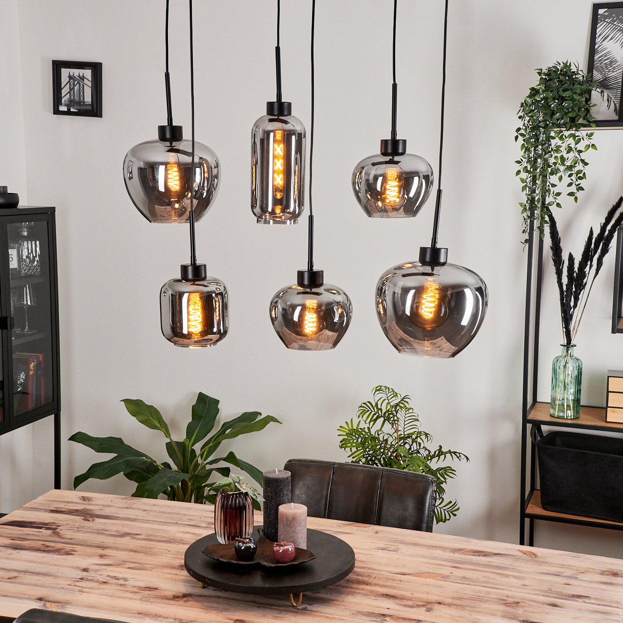 Ein Esszimmer mit sechs Pendelleuchten in verschiedenen Formen, mit leuchtenden Glühbirnen. Ein Holztisch und grüne Pflanzen.