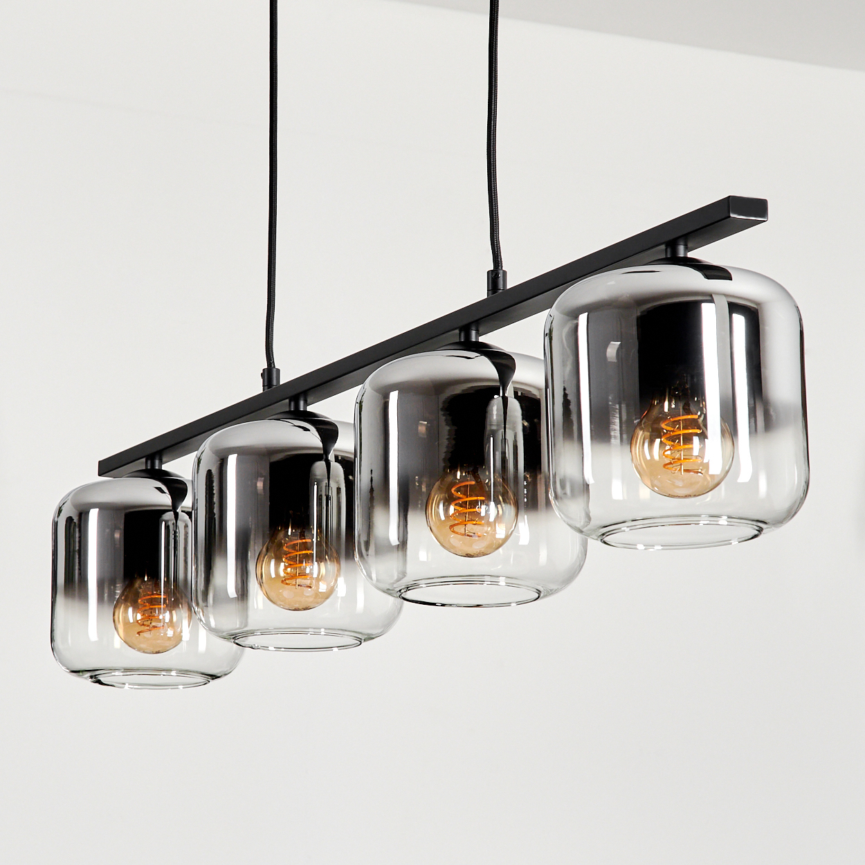 Eine moderne Deckenleuchte. Vier Glasschirme mit leuchtenden Glühbirnen hängen an einer schwarzen Metallstange. Der Hintergrund ist weiß.