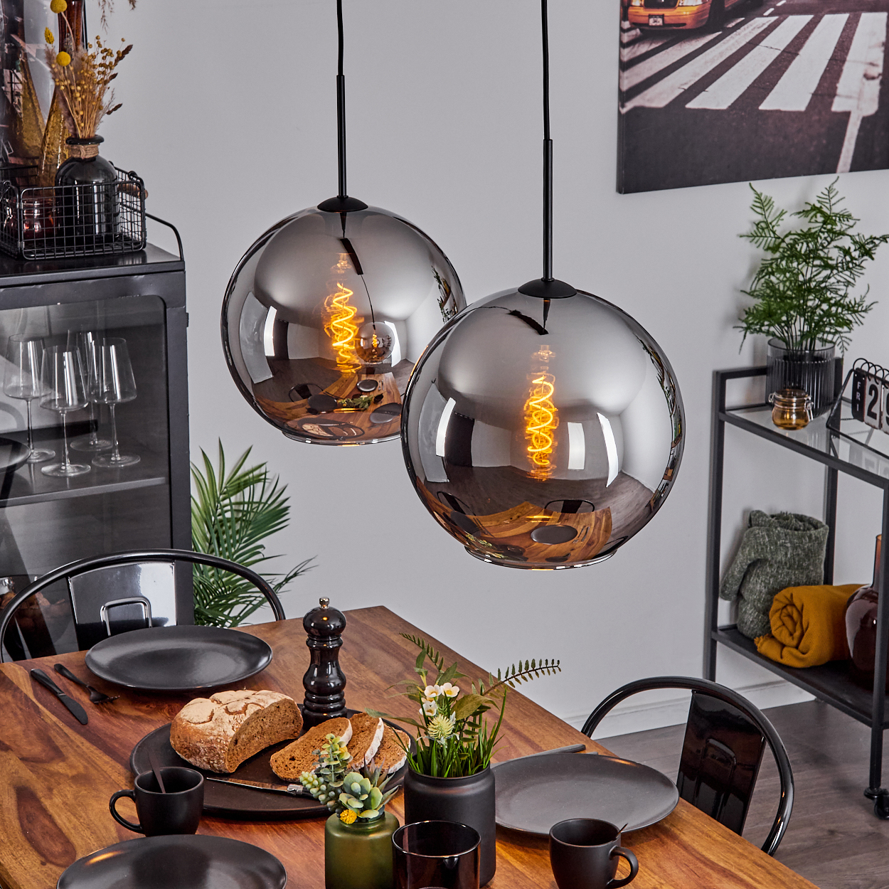 Zwei Pendelleuchten mit verspiegelten Glasschirmen hängen über einem Holztisch mit Essen und Deko.