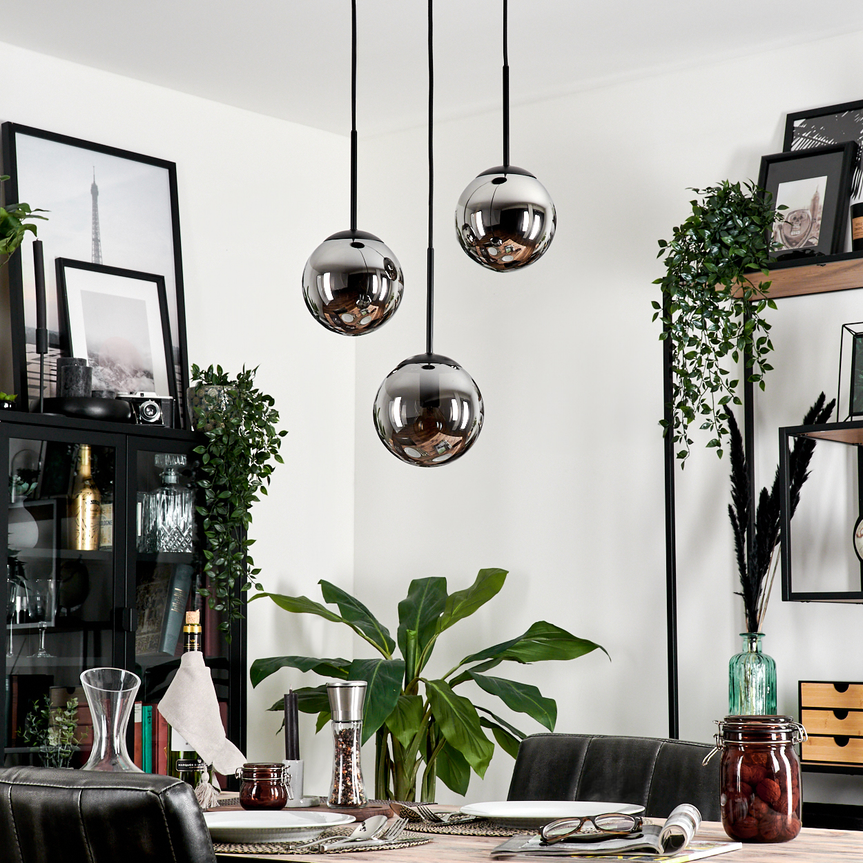 Esszimmer mit drei Pendelleuchten, jede mit einem rauchfarbenen Glaskugelschirm. Tisch gedeckt, Pflanzen und Kunstwerke sichtbar.