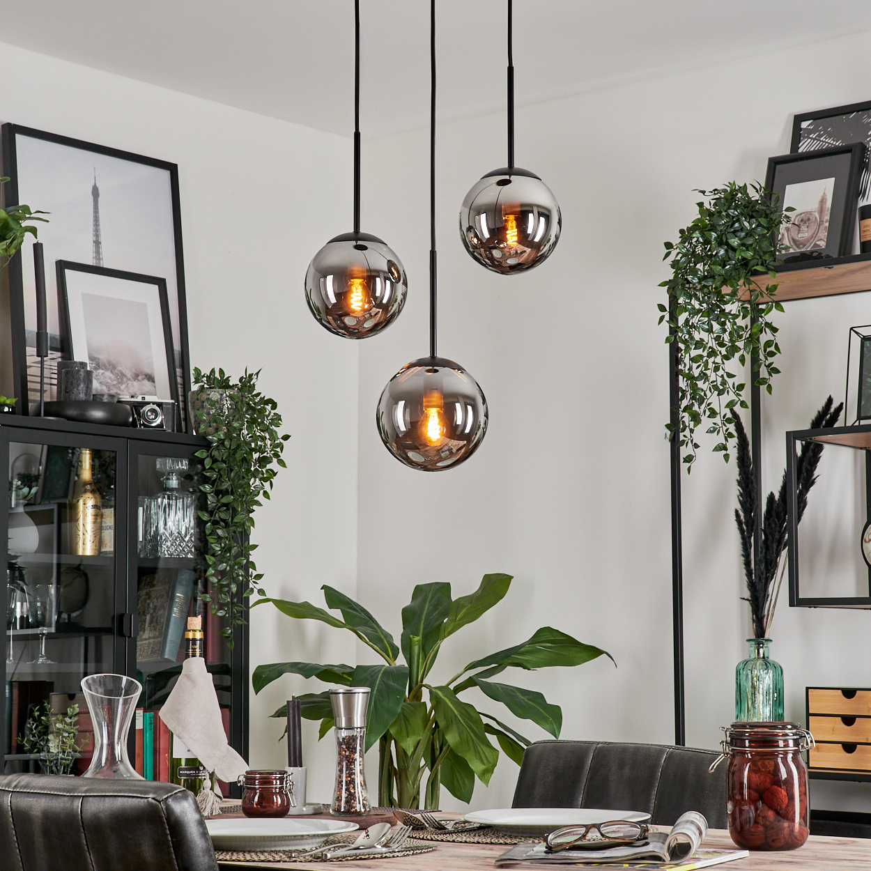 Esszimmer mit drei Pendelleuchten, jede mit einem rauchfarbenen Glaskugelschirm. Tisch gedeckt, Pflanzen und Kunstwerke sichtbar.