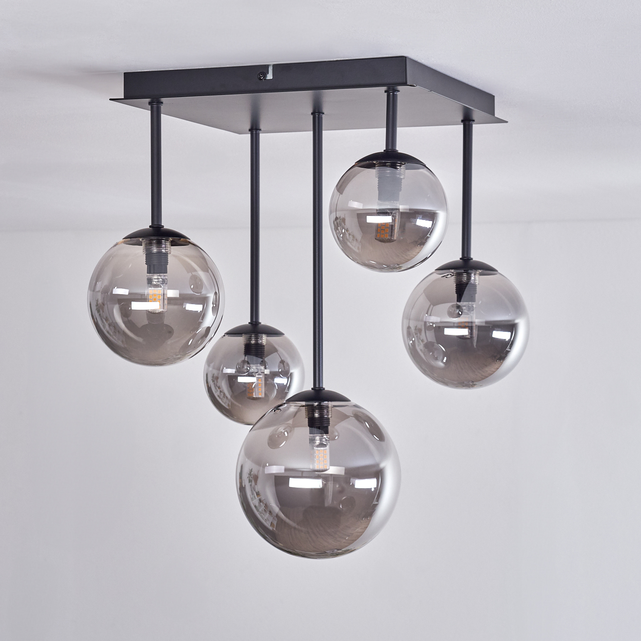 Deckenlampe mit fünf rauchigen Glaskugeln. Schwarzer quadratischer Sockel. Innenraum mit weißen Wänden. Die Lampen sind beleuchtet.