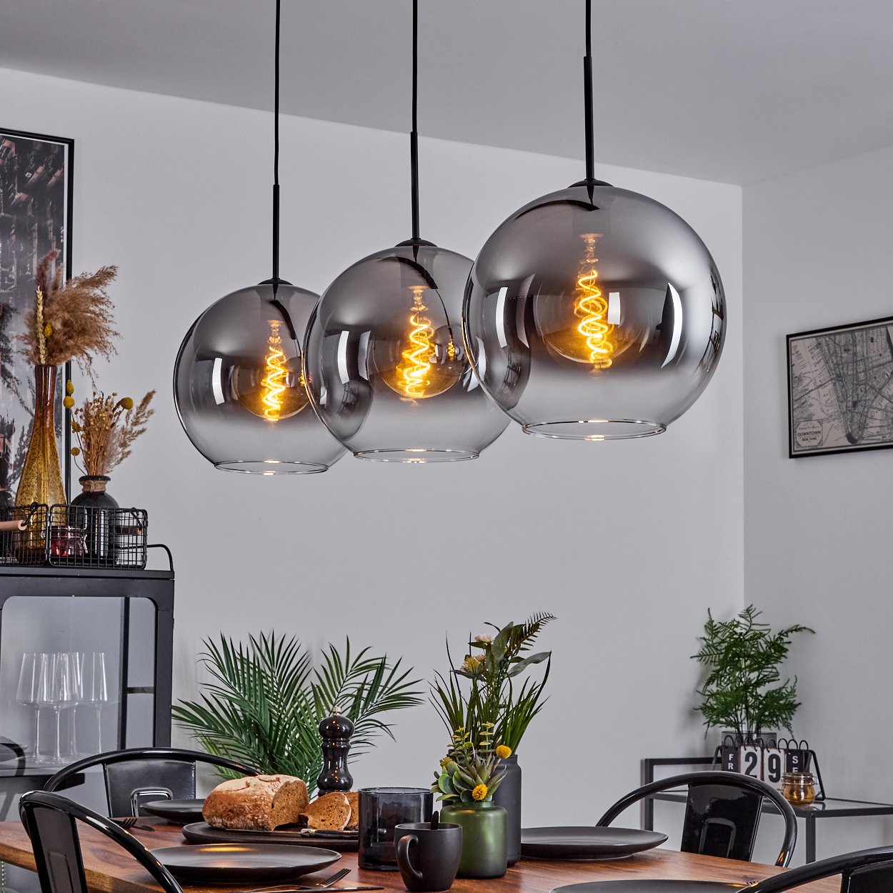 Drei kugelförmige Lampen hängen über einem Esstisch. Die Lampen haben Wendelglühbirnen. Der Raum hat schwarze Möbel.