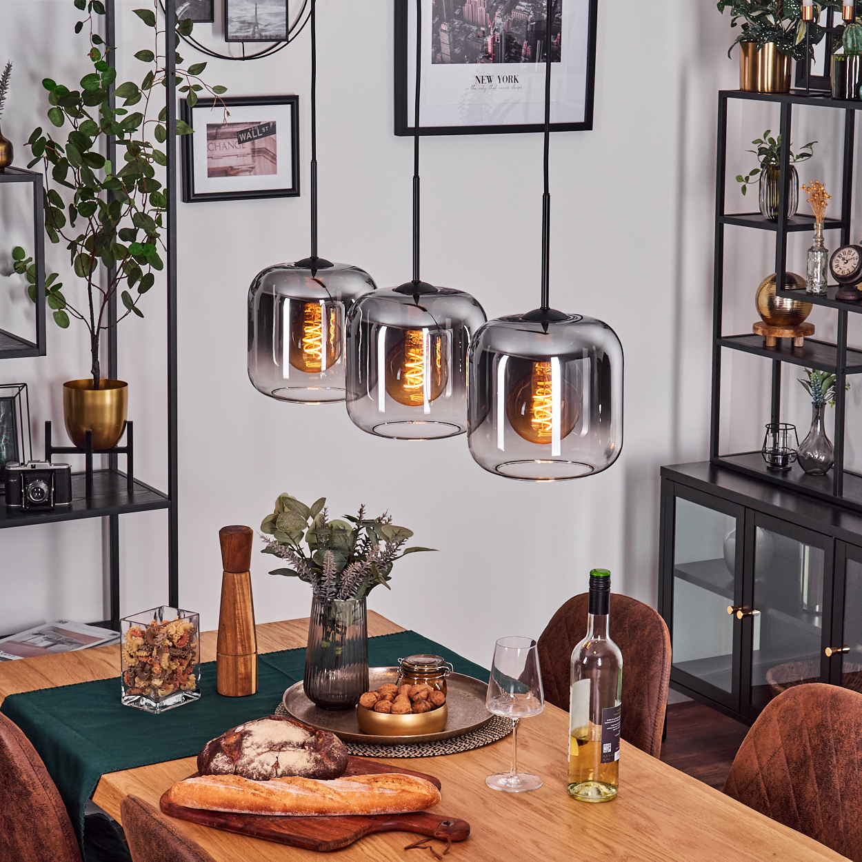 Drei Lampen mit rechteckigen Glasschirmen und sichtbaren Glühbirnen hängen über einem gedeckten Tisch. Pflanzen und Dekoration im Hintergrund.