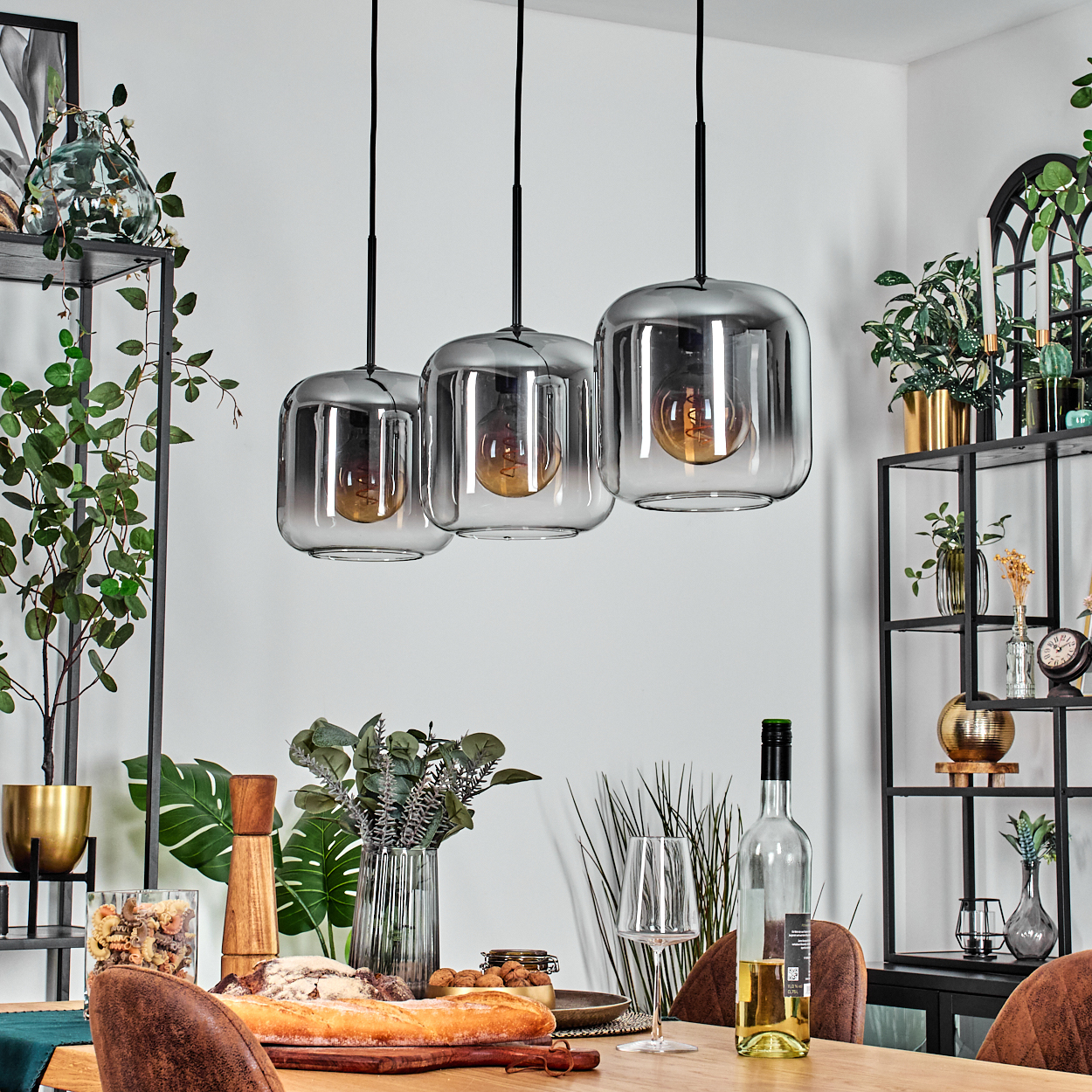 Drei Lampen mit rechteckigen Glasschirmen und sichtbaren Glühbirnen hängen über einem Tisch. Pflanzen und Dekoration sind im Hintergrund.
