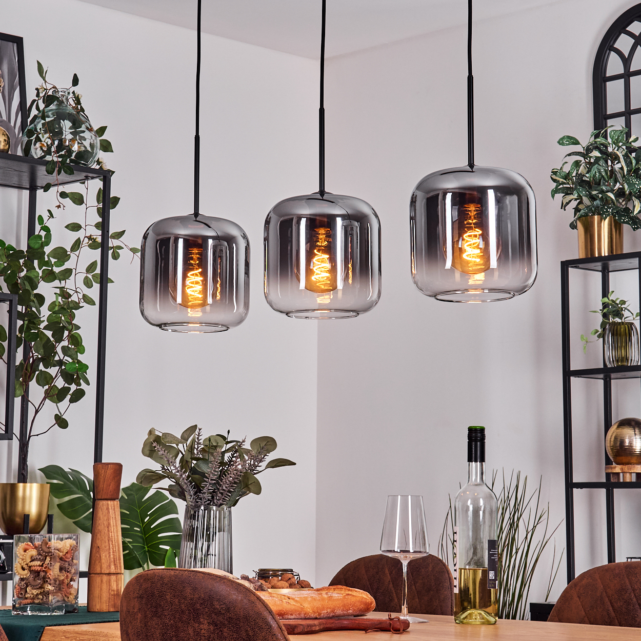 Drei Lampen mit rechteckigen Glasschirmen und sichtbaren Glühbirnen hängen vor einer weißen Wand. Pflanzen und Dekoration im Hintergrund.