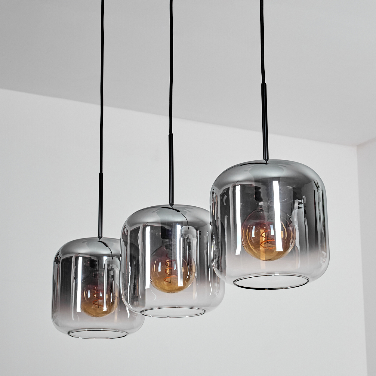 Drei Lampen mit rechteckigen Glasschirmen und sichtbaren Glühbirnen hängen an schwarzen Kabeln vor weißem Hintergrund.