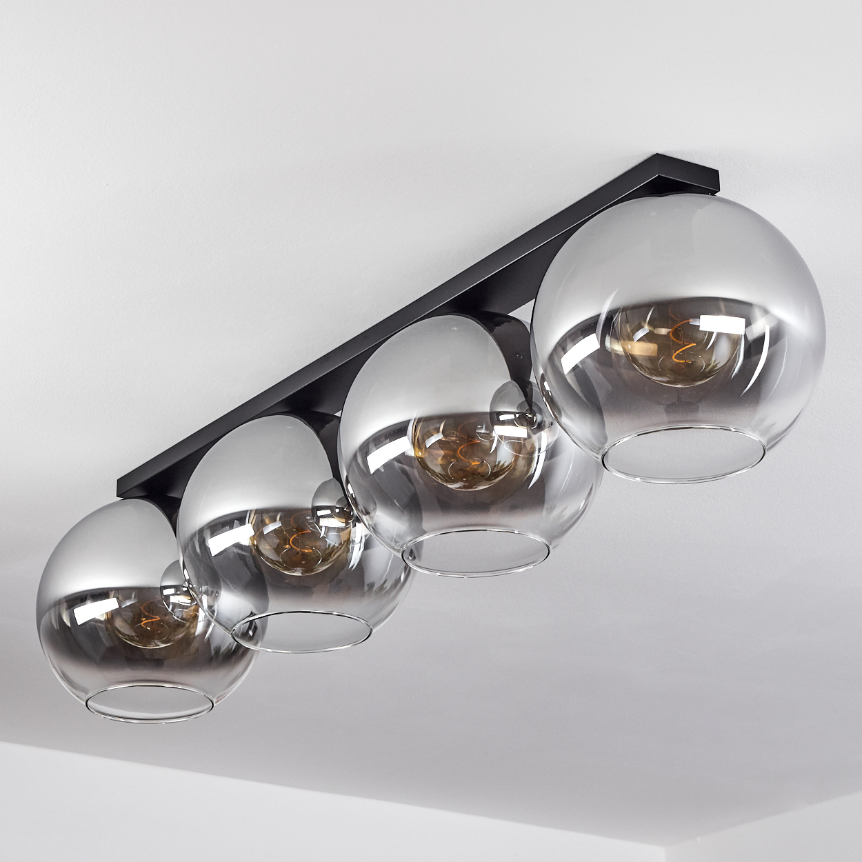 Deckenlampe mit vier kugelförmigen Schirmen. Jeder enthält eine Glühbirne. Die Schirme sind klar und der Hintergrund ist weiß.